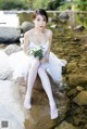 A woman in a white dress sitting on a rock by a river.