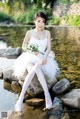 A woman in a white dress sitting on a rock by the water.