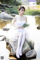 A woman in a white dress sitting on a rock by the water.